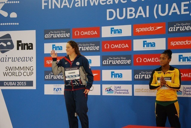 Molly Hannis waiving as she steps on the podium, with Alia Atkinson looking on.